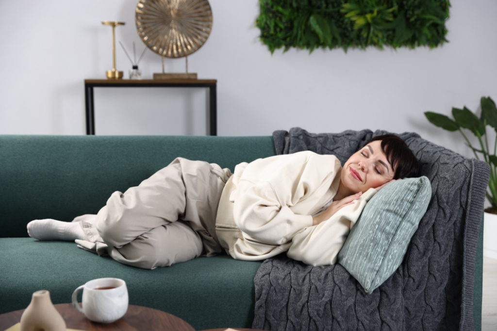 Nainen nukkuu kylkiasennossa vihreällä sohvalla pää tyynyä vasten, viltti ympärillään ja kahvikuppi pöydällä, kodikkaassa ja rauhallisessa olohuoneessa.