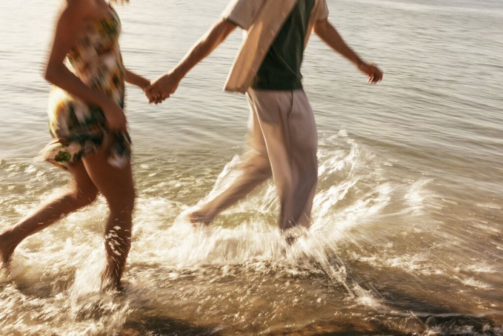 Kaksi aikuista juoksee käsi kädessä rantavedessä. 