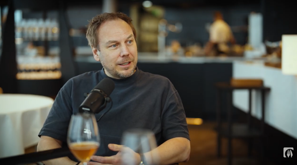 Keittiömestari Tommi Tuominen istuu mikrofoni edessä ravintolasalissa lasi edessä.