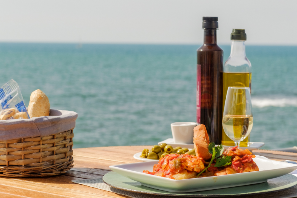 Rantaterassilla katettu pöytä meren äärellä: lautasella tomaattinen kala-annos ja leipää, vieressä oliiviöljy- ja viinipullo, lasi valkoviiniä sekä kori leipää, taustalla avautuu sininen meri.