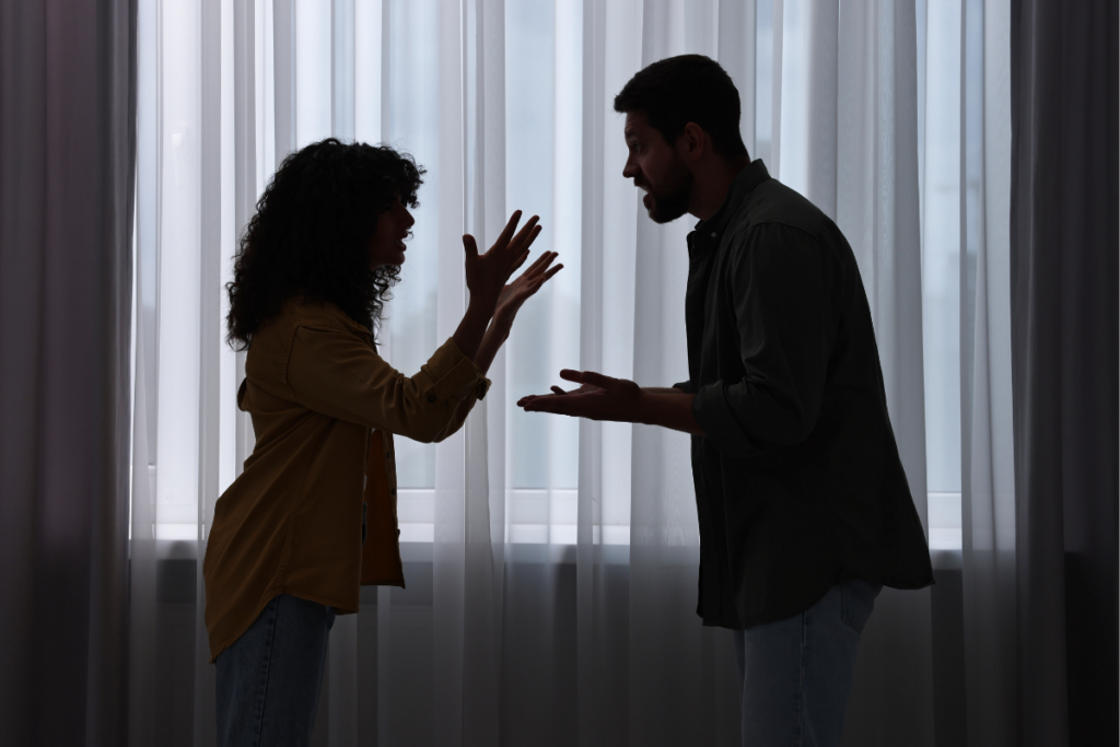 Kaksi aikuista keskustelee kiivaasti toisiaan vasten seisten, molemmat elehtivät käsillään. Taustalla oleva valo ja varjot vahvistavat tunteikasta ja konfliktista ilmapiiriä.