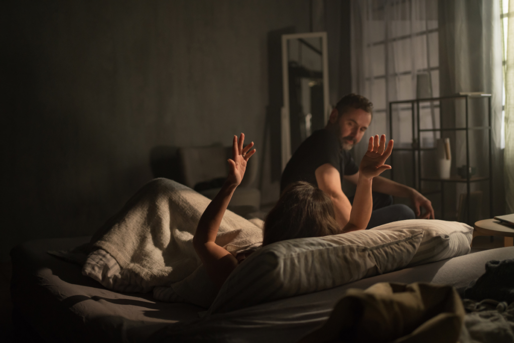 Hämärässä makuuhuoneessa pariskunta on ristiriitatilanteessa: toinen makaa sängyssä kädet koholla puolustavasti, toinen istuu vieressä ja katsoo häntä etäältä, tunnelma on jännittynyt ja riitaisa.