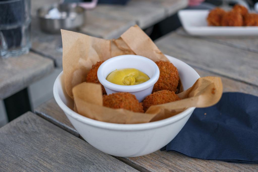 Friteerattuja pyöryköitä tai kroketin kaltaisia palleroita, jotka on tarjoiltu kulhossa ruskean paperin päällä, keskellä kirkkaankeltainen sinappidippi.