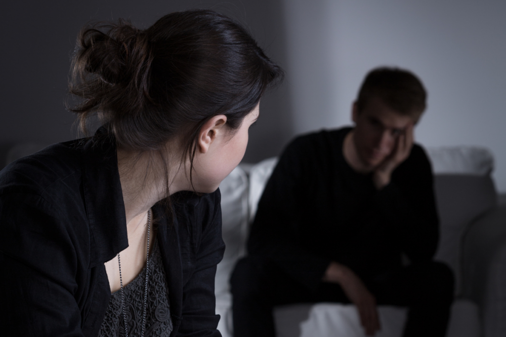 Etu-alalla nainen katsoo huolestuneena miestä, joka istuu sohvalla käsi kasvoillaan; molempien olemus on etäinen ja surullinen, mikä kuvastaa vaikeaa tilannetta parisuhteessa.