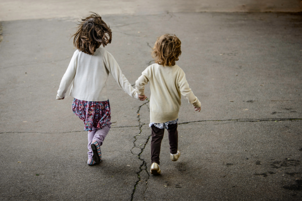 Kaksi pientä lasta kävelee käsi kädessä poispäin kamerasta asfaltilla, toinen hieman toista pidempi. Heillä on vaaleat neuleet ja liike näyttää huolettomalta.
