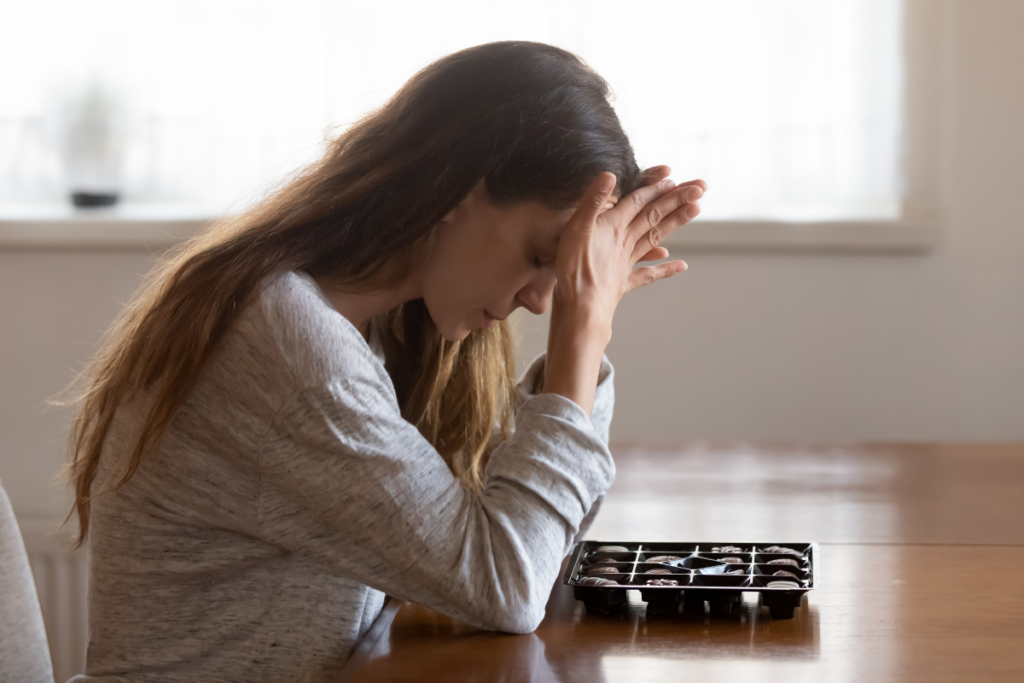 Nainen istuu pöydän ääressä sisätiloissa pää kumartunutena ja käsi otsalla. Hänen edessään pöydällä on avoin suklaarasia, jossa näkyy useita konvehteja.