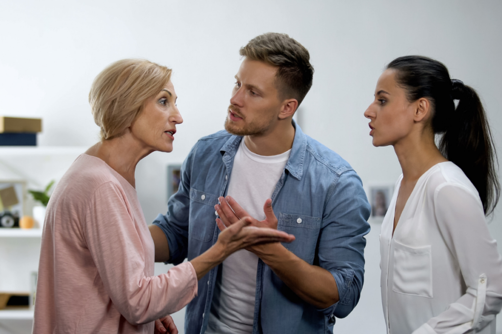 Kolme aikuista sisätilassa riitelemässä: vanhempi nainen puhuu kiihtyneesti, mies yrittää rauhoitella elehtimällä käsillään ja nuorempi nainen seisoo sivussa jännittyneenä ja vastaa sanallisesti.