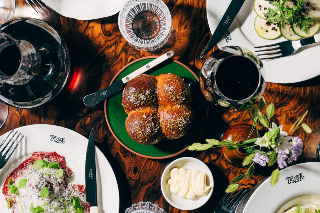 Ravintolapöytä ylhäältä kuvattuna: briossisämpylöitä, alkuruokia ja viinilaseja.