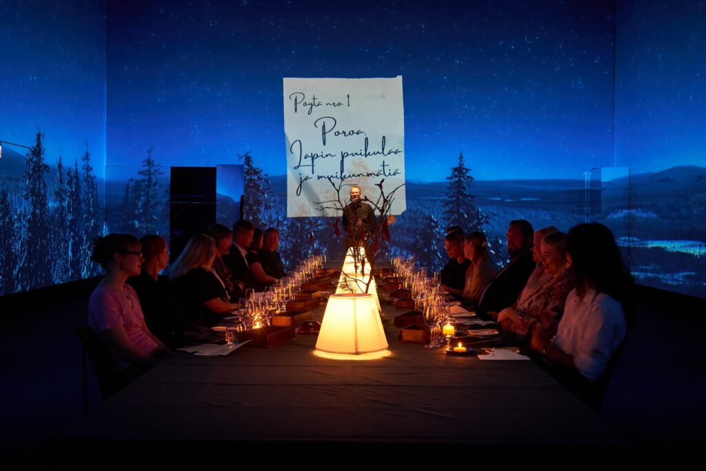 Pitkän pöydän ääressä istuvia illallisvieraita elämyksellisessä ravintolatapahtumassa Logomossa.