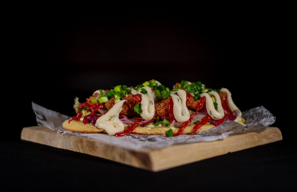 Pitkulainen leipäannos, jossa friteerattua kanaa, kastikkeita ja kevätsipulia puualustalla.