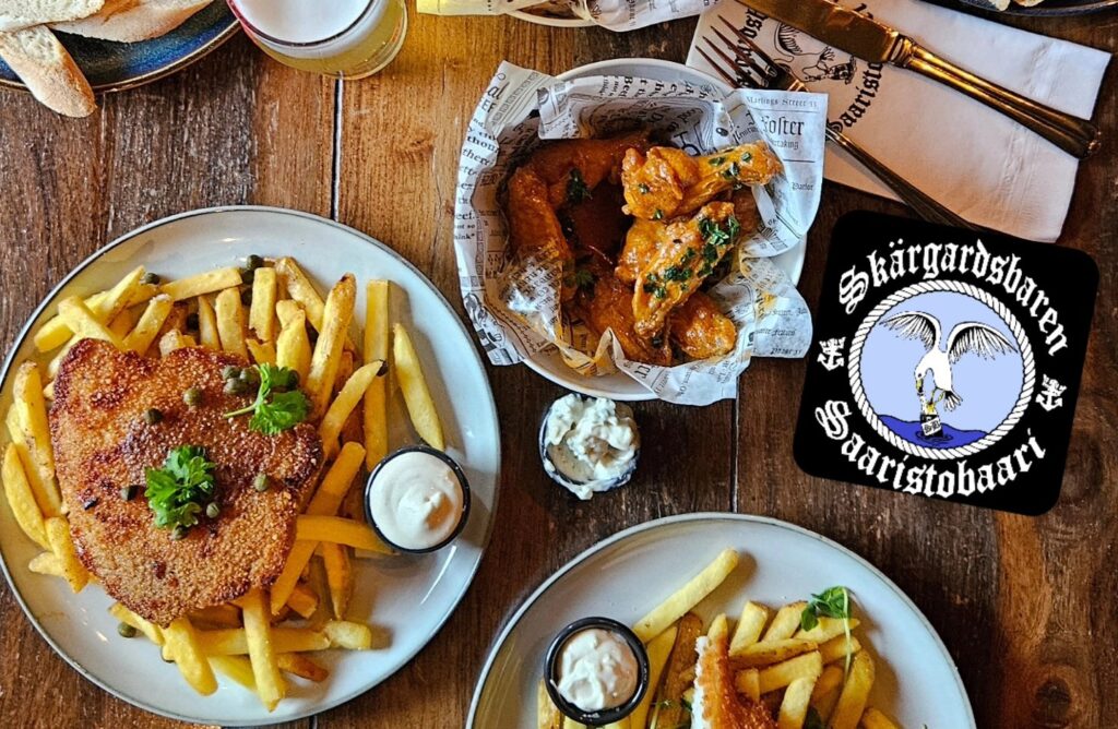 Pöydällä annoksia: leivitetty schnitzel ranskalaisten ja kastikkeen kanssa, kulhollinen kanansiipiä sekä dippejä.