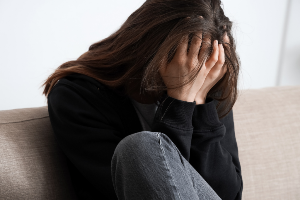 Nuori nainen istuu sohvalla polvet koukussa ja peittää kasvonsa molemmilla käsillä. Hänen kehonkielensä ja asennon perusteella hän vaikuttaa ahdistuneelta tai voimakkaasti surulliselta.