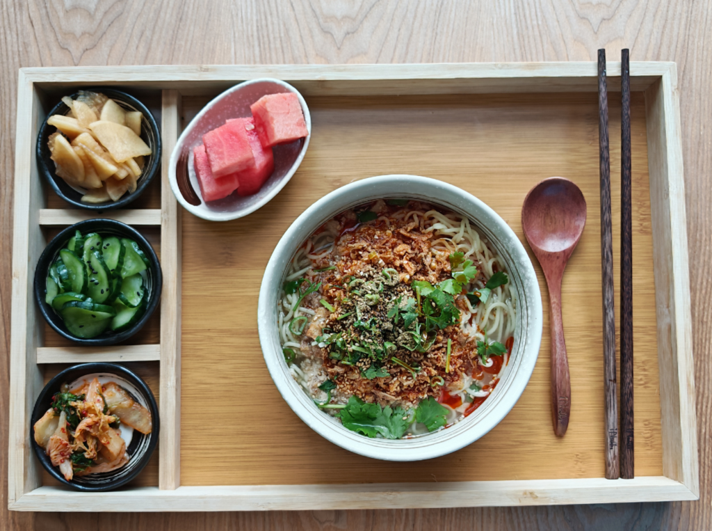 Nuudelikeitto tarjoilualustalla, lisukkeina pikkukulhot vihanneksia ja vesimelonia sekä syömäpuikot.