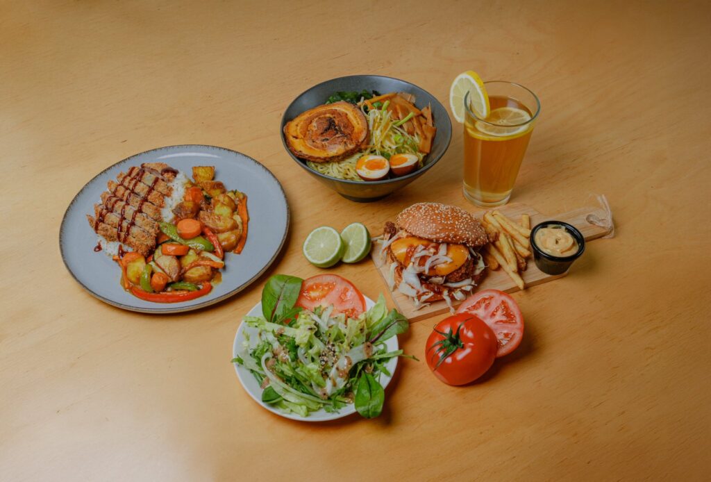 Pöydälle katettu useita ravintola-annoksia, mukana ramenkulho, hampurilainen ranskalaisilla ja dipillä, vihersalaatti sekä riisi- ja kasvisannos.