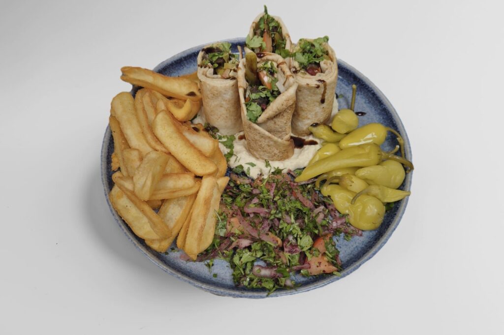 Lautasellinen Lähi-idän ruokaa: shawarma-rullia, ranskalaisia perunoita, hummusta, salaattia ja säilöttyjä vihreitä chilejä.