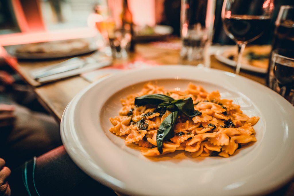 Pastaruoka valkoisella lautasella ravintolapöydässä, taustalla viinilaseja ja muita annoksia