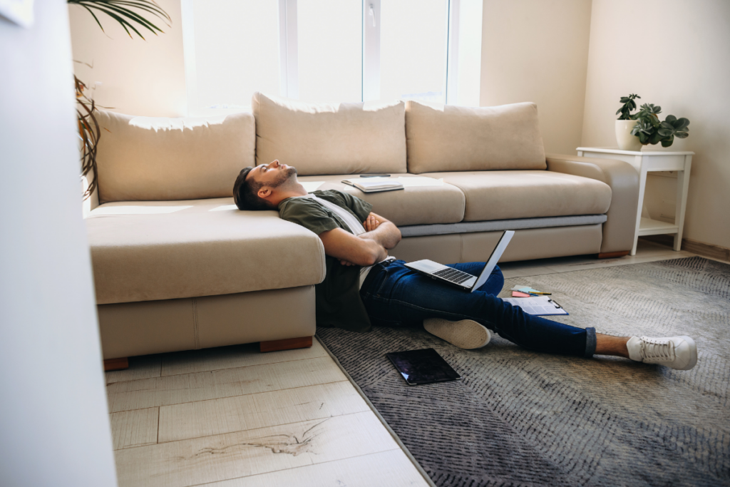 Mies makaa olohuoneen lattialla nojaten sohvaan, silmät kiinni ja kädet ristissä, läppäri sylissä ja työvälineitä ympärillä, selvästi uupuneena työpäivän jälkeen.