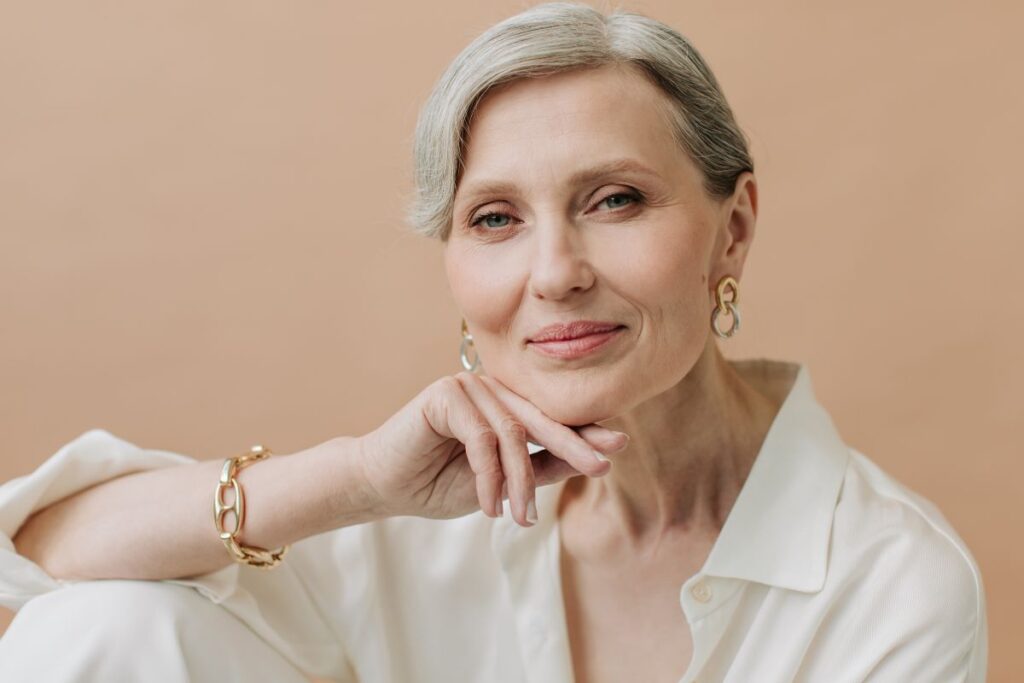 Aikuinen nainen vaaleassa asussa katsoo kameraan ja nojaa leukaa käteensä vaaleaa taustaa vasten.