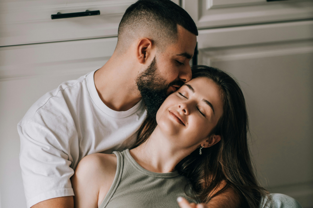 Mies suutelee hellästi naista ohimolle; nainen hymyilee silmät suljettuina ja nojaa mieheen, mikä kuvastaa turvallisuutta, luottamusta ja emotionaalista läheisyyttä.