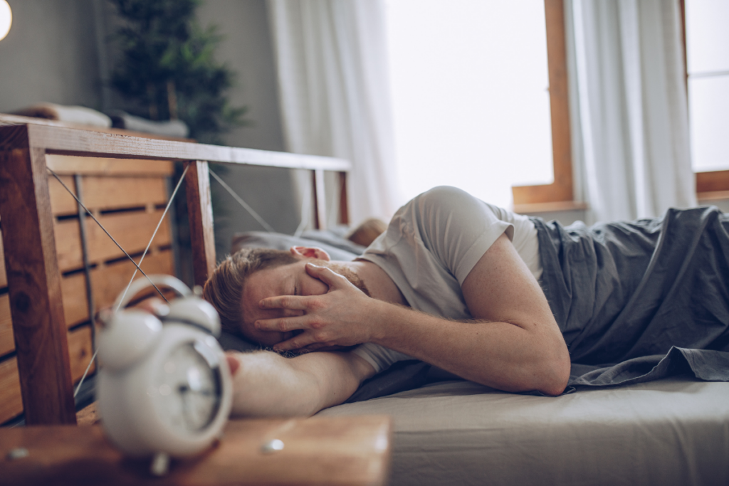 Nuori mies makaa sängyssä herätyskellon vieressä peittäen kasvonsa kädellään, ilme viestii väsymystä ja vaikeaa heräämistä aamulla.