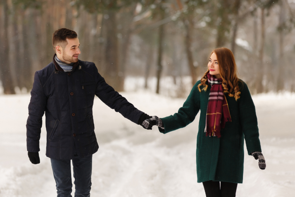 Nuori mies ja nainen kävelevät käsi kädessä lumisessa metsämaisemassa, hymyilevät toisilleen ja ovat pukeutuneet lämpimiin takkeihin, huiveihin ja hansikkaisiin.