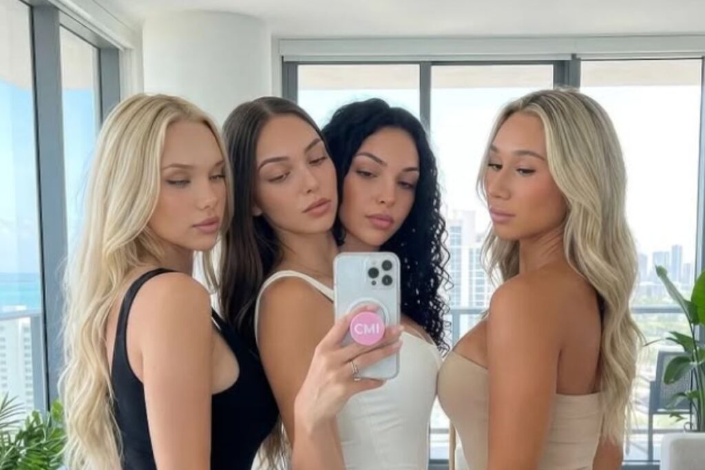 Neljä nuorta naista poseeraa peiliselfiessä valoisassa asunnossa, taustalla suuret ikkunat ja kaupunkimaisema