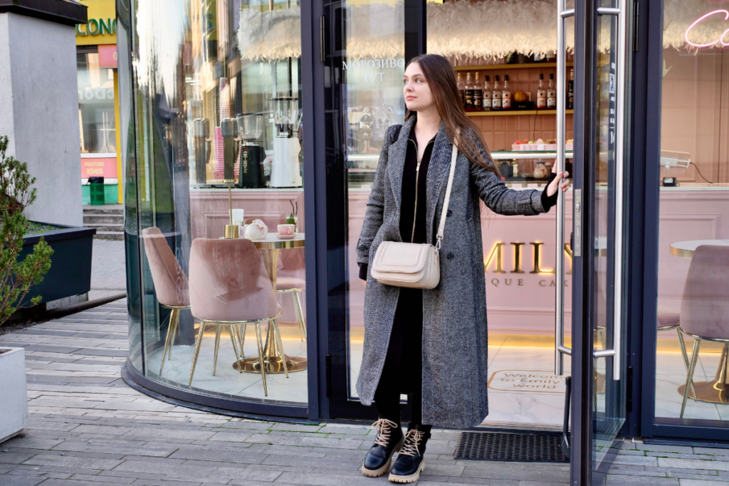 Sama nainen astuu ulos vaaleanpunasävyisestä kahvilasta, pitää lasiovea auki ja katsoo kadulle; hänellä on pitkä harmaa takki, mustat vaatteet, nilkkurit ja beige olkalaukku.