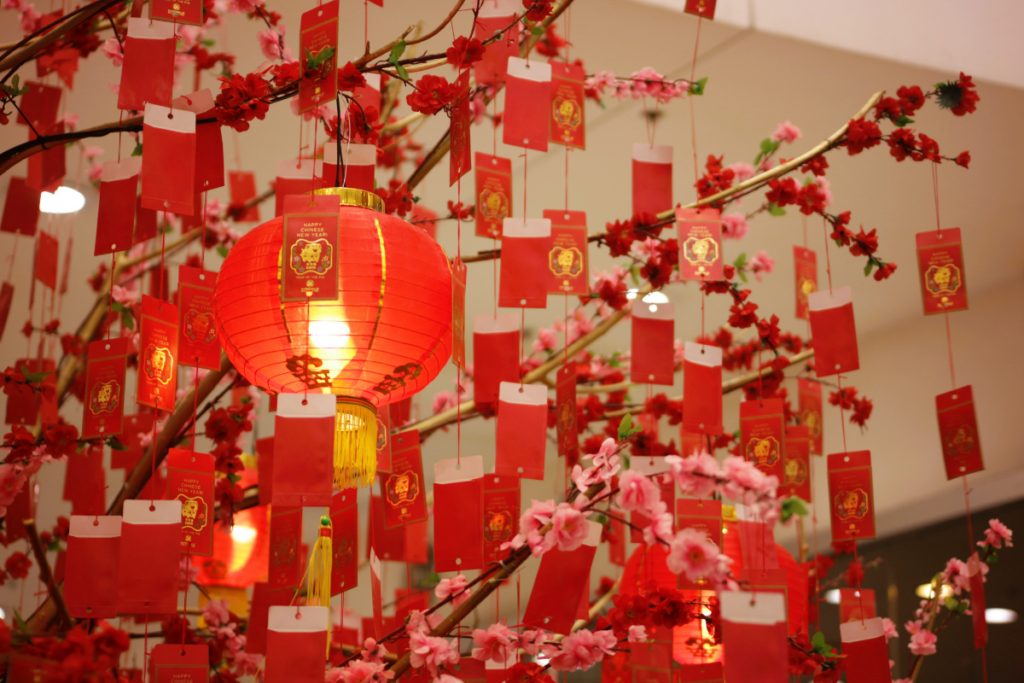 Punainen kiinalainen lyhty ja kukkiva koristepuu, johon on ripustettu punaisia uudenvuoden toivotuskortteja.