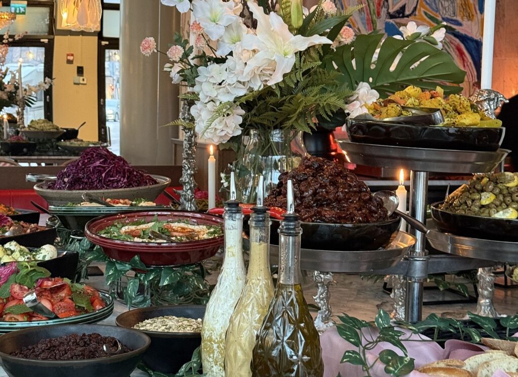 Runsas buffetpöytä ravintola Biblosissa: värikkäitä salaatteja, lämpimiä kasvisruokia, kastikkeita ja leipiä esillä koristeellisesti kynttilöiden ja kukka-asetelman ympäröimänä.