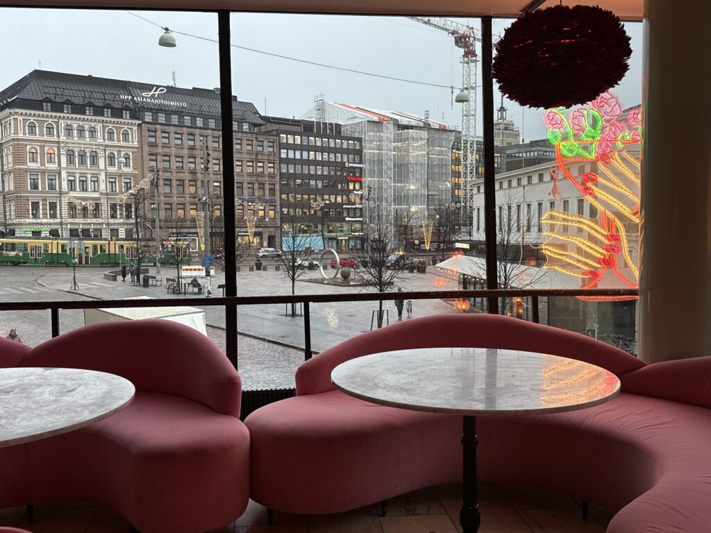 Ravintolan vaaleanpunaiset nojatuolit ja pyöreät pöydät suurten ikkunoiden äärellä, näkymä kaupunkiaukiolle.