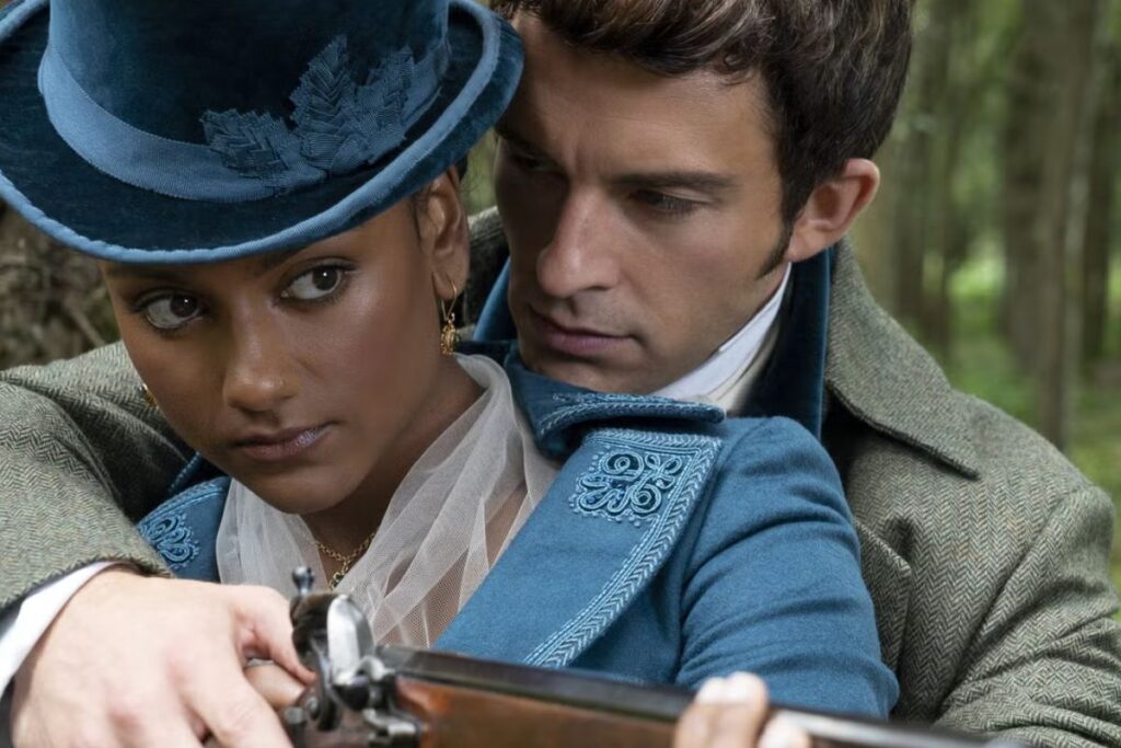 Nuori nainen sinisessä hatussa ja takissa pitää käsissään vanhanaikaista asetta, kun mies hänen takanaan ohjaa häntä läheltä metsäisessä ympäristössä.