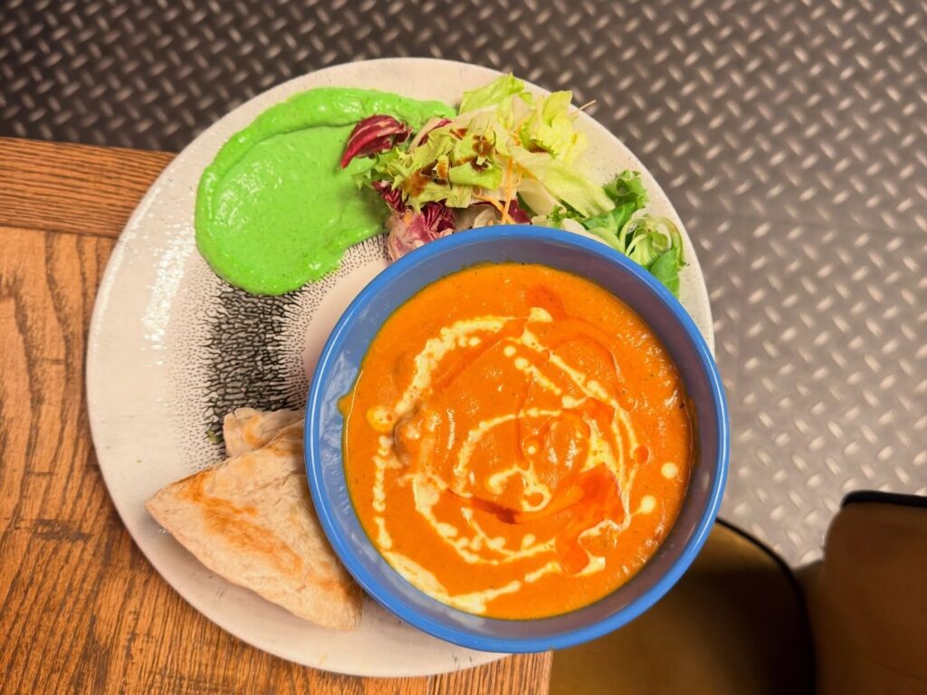 Ylhäältä kuvattu annos: oranssia curryä sinisessä kulhossa, vihreää kastiketta, vihreää salaattia ja pala naan-leipää vaalealla lautasella puupöydällä.