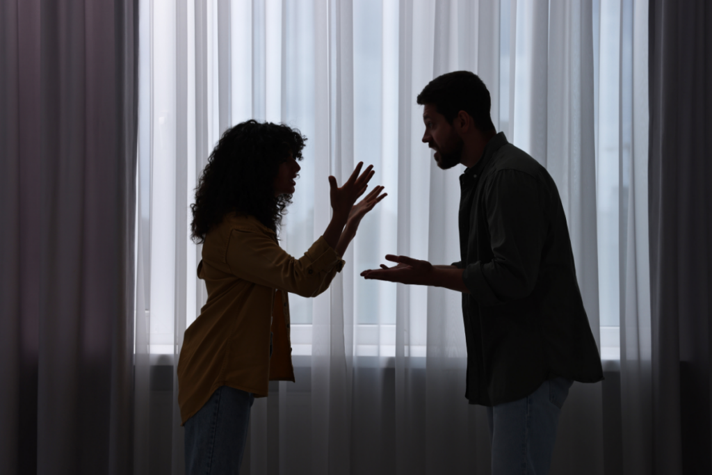 Mies ja nainen riitelevät vastakkain ikkunan edessä; molemmat elehtivät käsillään hämärässä huoneessa.