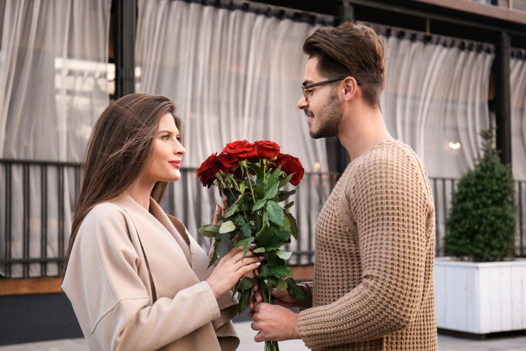 Mies antaa naiselle punaisista ruusuista koostuvan kukkakimpun ulkona, ja he katsovat toisiaan hymyillen.