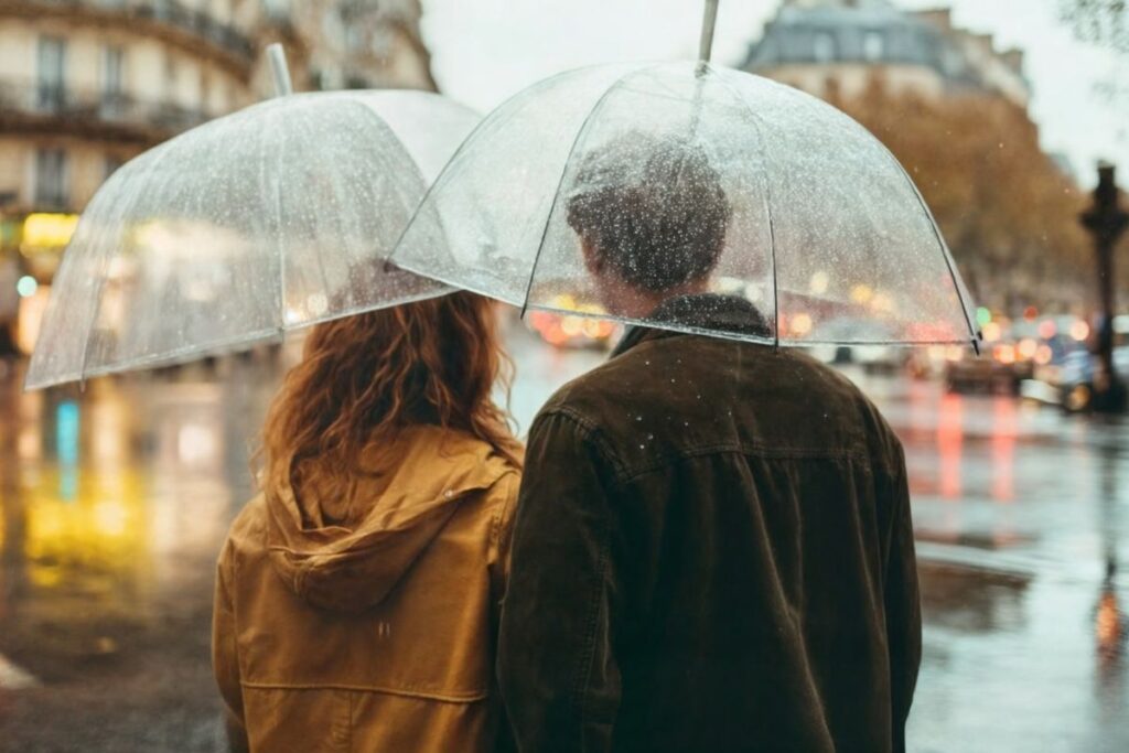 Pari seisoo urbaanissa ympäristössä sateella läpinäkyvien sateenvarjojen kanssa. Sateenvarjoissa näkyy pisaroita ja pariskunnalla on säänmukainen varustus.