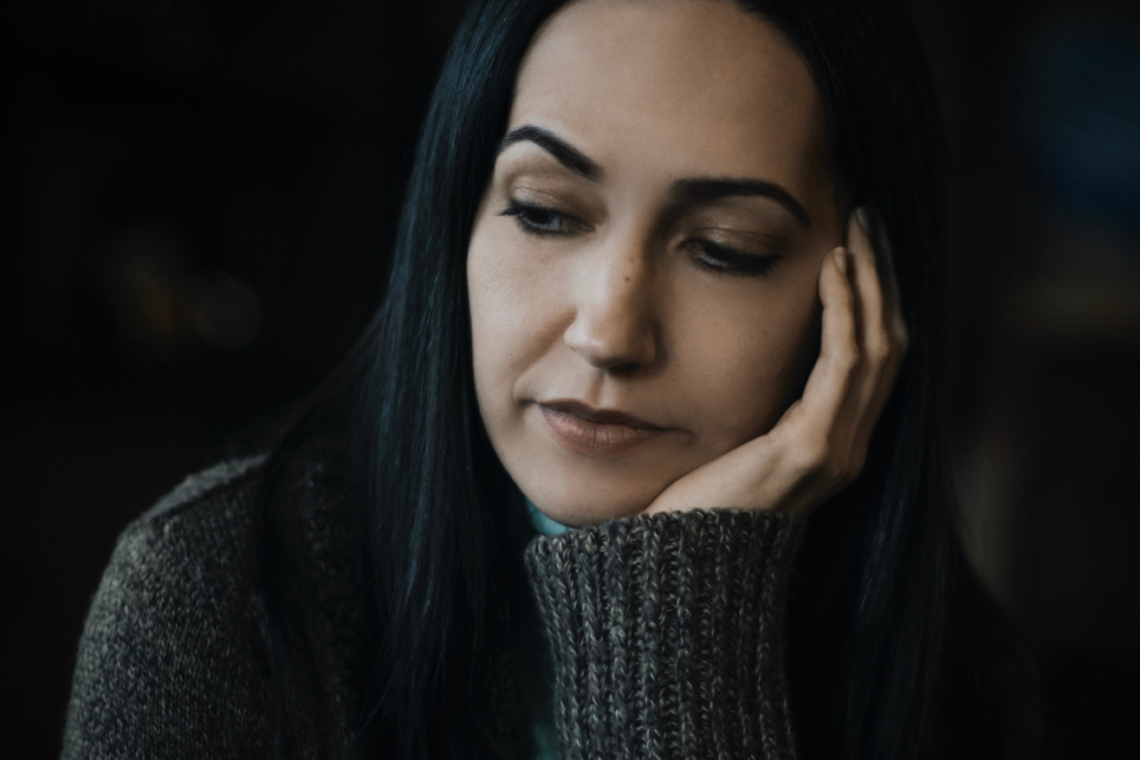 Nainen katsoo alaspäin mietteliäänä ja nojaa poskeaan käteensä hämärässä valaistuksessa.