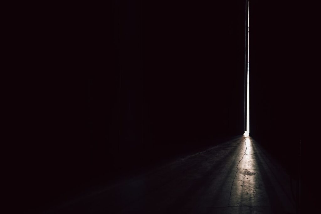Pimeä käytävä tai huone, jonka päässä näkyy kapea valojuova ovenraosta.