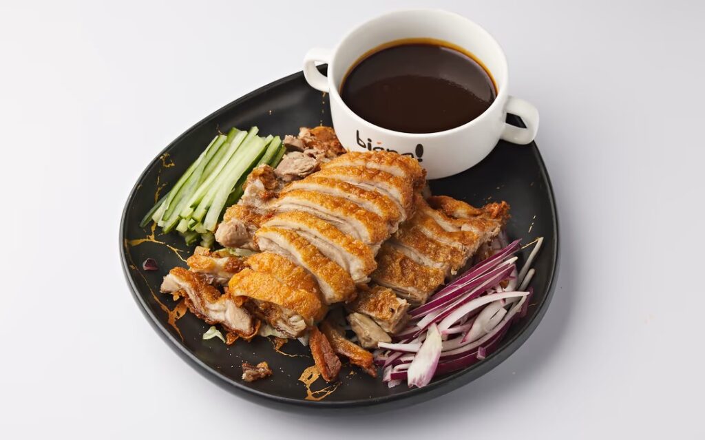 Viipaloitua rapeakuorista ankkaa lautasella, vierellä kurkkusuikaleita, punasipulia ja kulhollinen tummaa kastiketta.