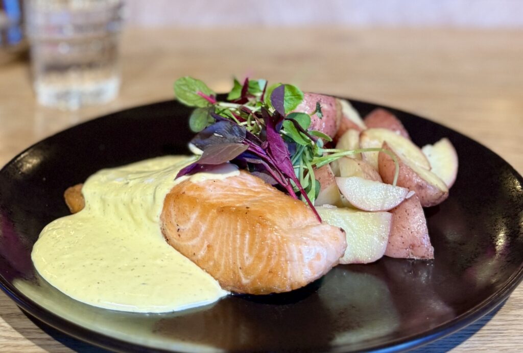 Paistettua lohta keltaisen kastikkeen kanssa, lisukkeena perunoita ja tuoreita versoja mustalla lautasella.