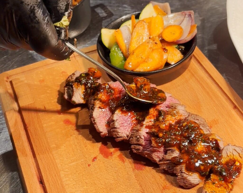 Viipaleiksi leikattu liha leikkuulaudan päällä. Päälle asetellaan maustevoita ja vieressä kasviksia kulhossa.