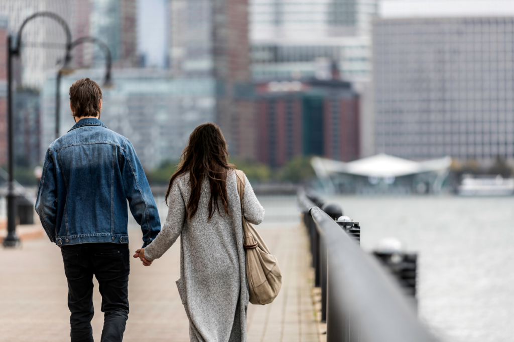 Kaksi aikuista henkilöä kävelee käsi kädessä rantapromenadilla, taustalla kaupunkimaisema