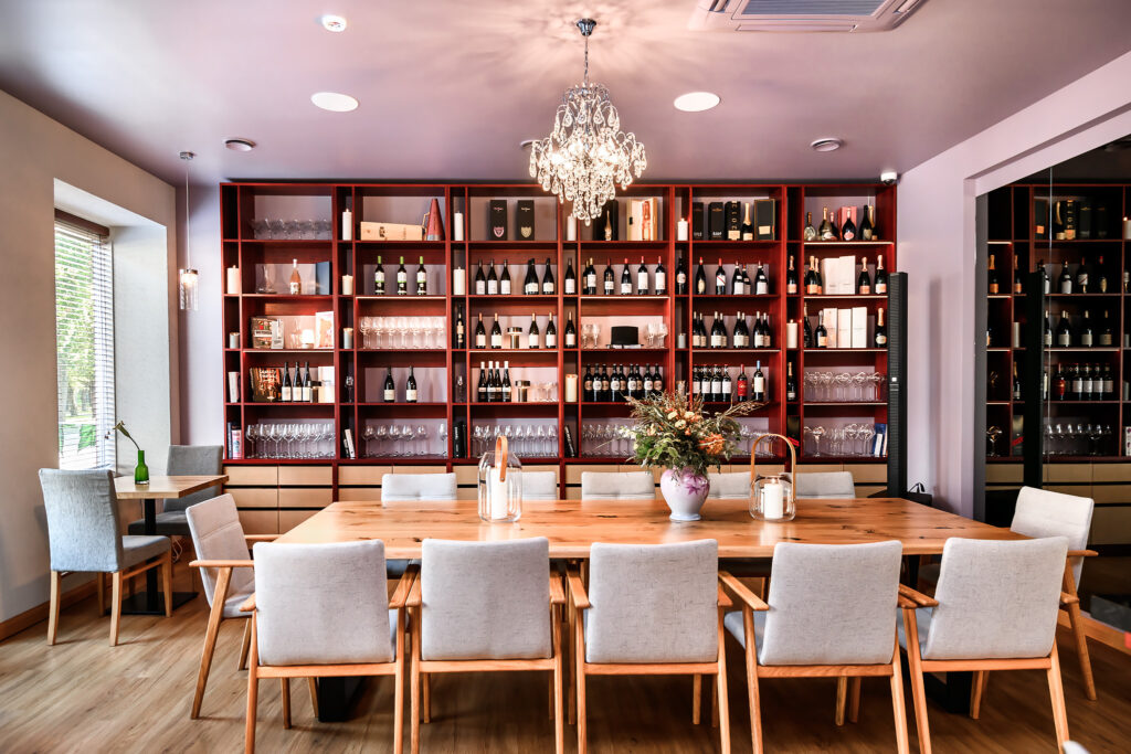Ravintolan tyylikäs sisätila: pitkä puinen pöytä, vaaleat tuolit ja seinällinen viinipulloja hyllyissä.