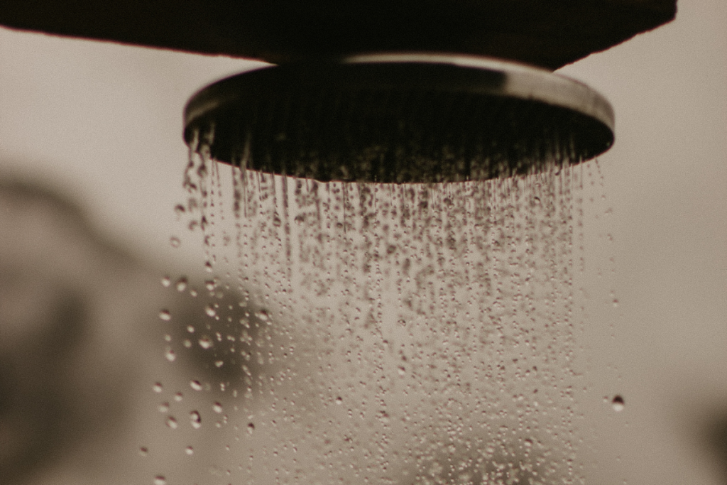 Lähikuva suihkupäästä, josta vesi virtaa tasaisena suihkuna vaaleaa taustaa vasten.