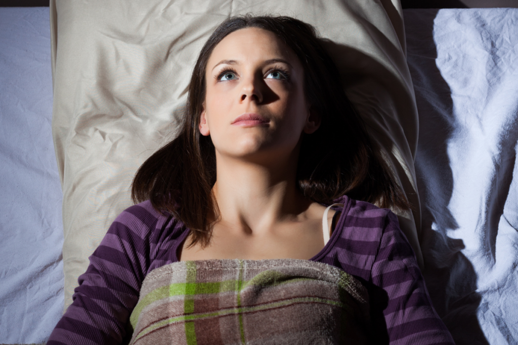 Aikuinen henkilö makaa selällään sängyssä peiton alla ja katsoo ylöspäin