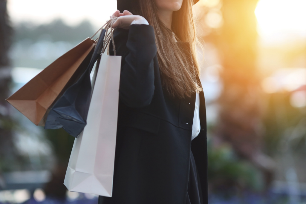 Nainen kävelee ulkona auringonlaskun valossa useita ostoskasseja olallaan, pukeutuneena tummaan takkiin.