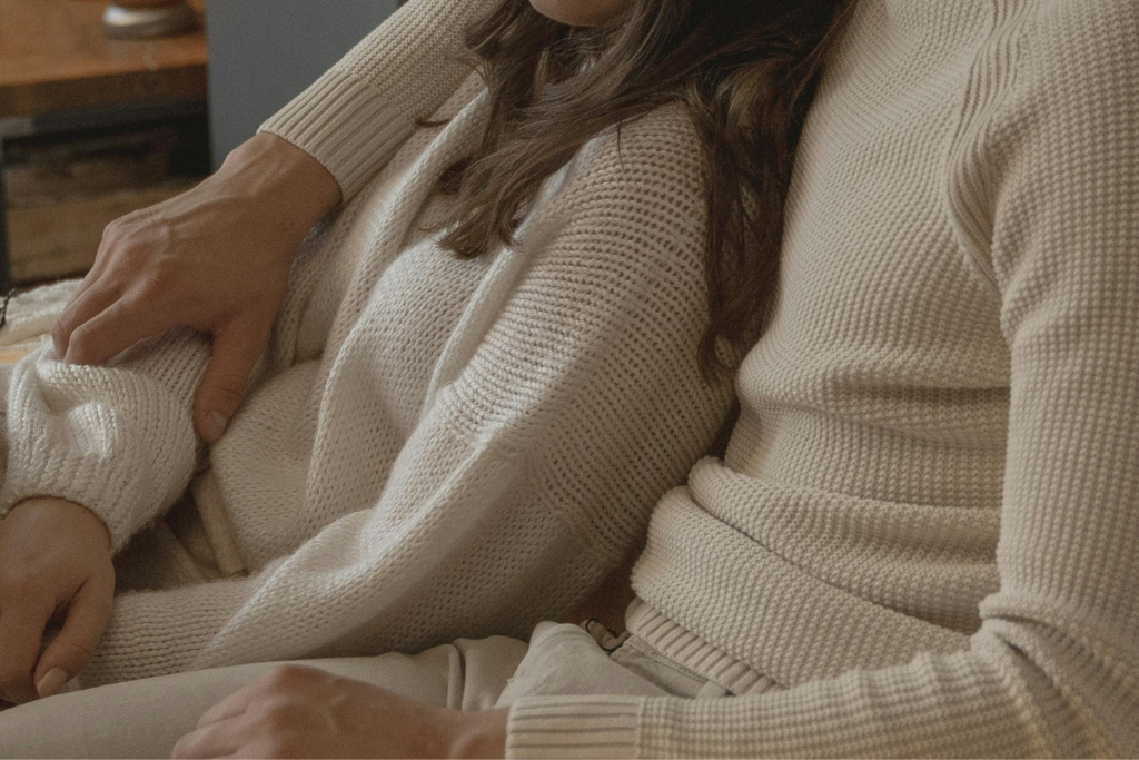 Pariskunta istuu lähekkäin sohvalla, toinen pitää kättään toisen sylissä rennossa ja läheisessä tunnelmassa.