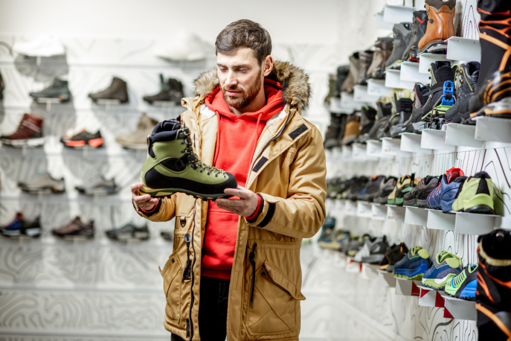 Mies tarkastelee vaelluskenkää urheilukaupassa, ympärillään esillä erilaisia kenkiä hyllyillä.