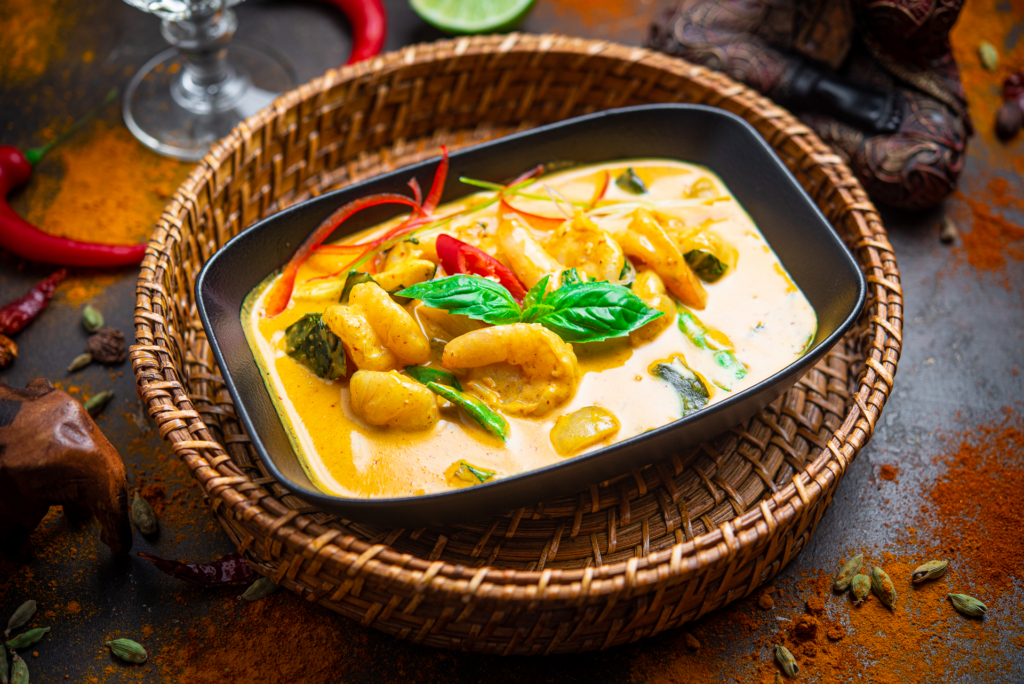 Thaimaalainen curryannos, jossa katkarapuja ja vihanneksia keltaisessa kookoskastikkeessa mustassa astiassa punotun korin päällä.