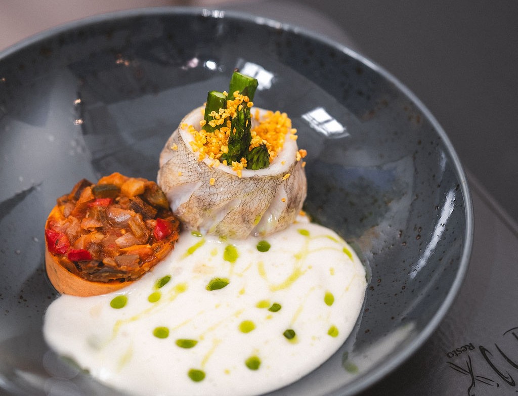 Tyylikkäästi aseteltu kala-annos vaalealla kastikkeella, vihreillä parsapaloilla ja kasvistartarilla harmaalla lautasella.