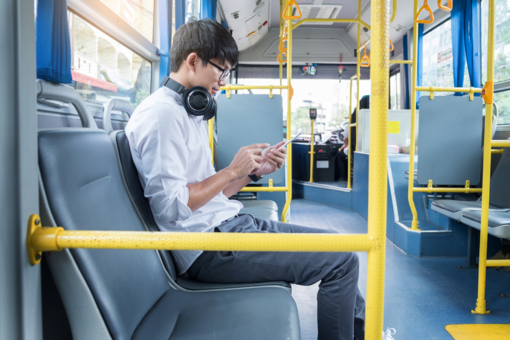 Kuulokkeita käyttävä nuori mies istuu bussin penkillä ja katsoo älypuhelintaan julkisessa liikenteessä.